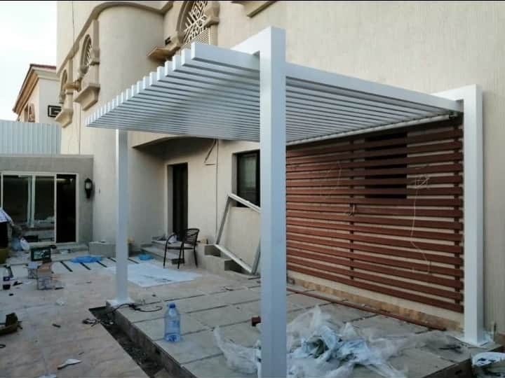 برجولا بشرائح ألمنيوم بيضاء مع ساتر خشبي في فناء منزل من تنفيذ عالم الظل الأول